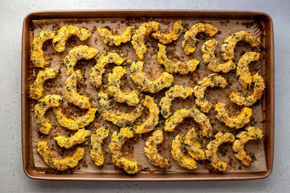Parmesan Crusted Roasted Delicata Squash arranged on a baking sheet lined with brown parchment paper. The baking sheet sits atop a creamy white textured surface.