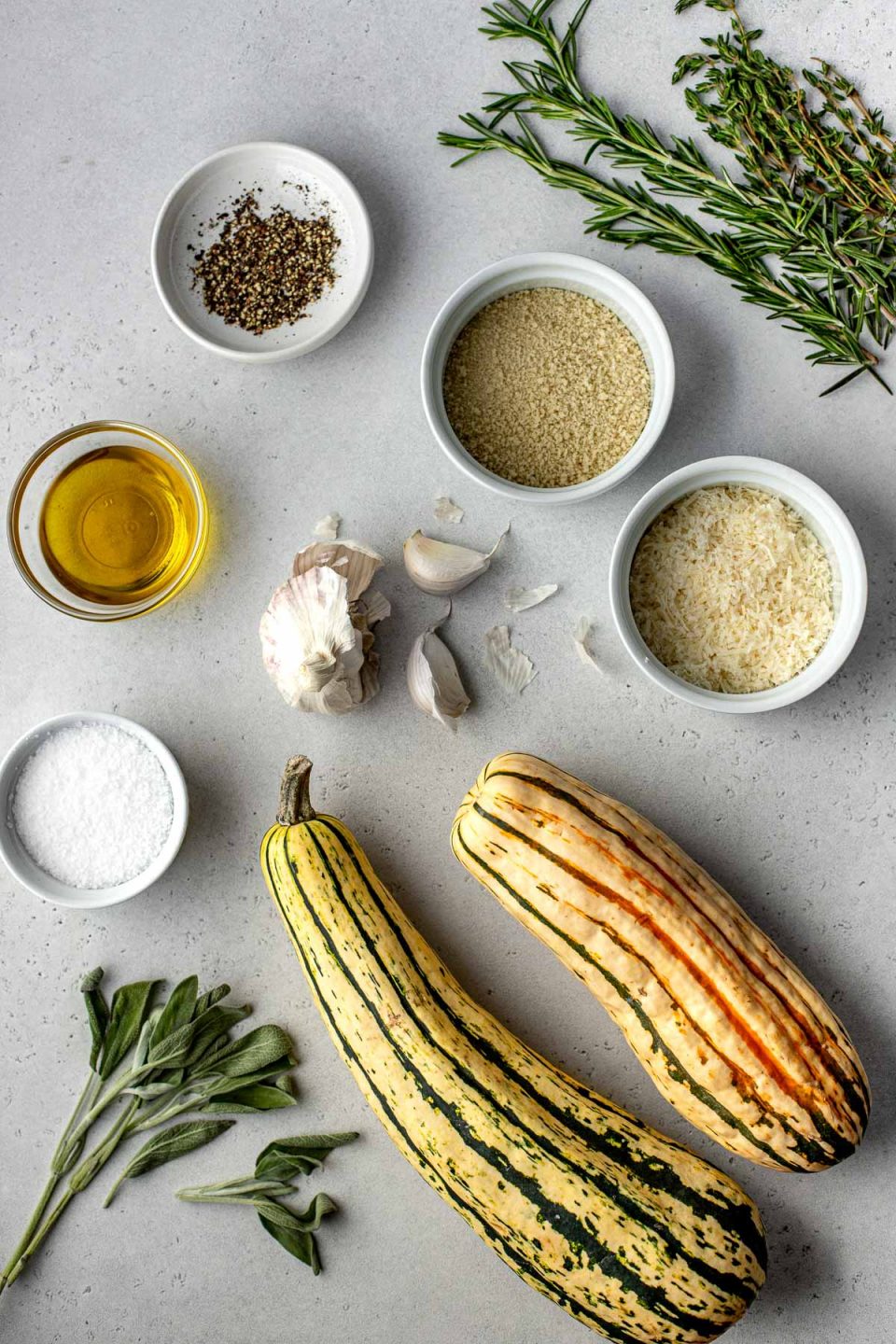 Parmesan Crusted Roasted Delicata Squash ingredients arranged on a creamy white textured surface: delicata squash, garlic, fresh herbs - rosemary, sage, and thyme, olive oil, kosher salt, ground black pepper, panko breadcrumbs, and parmesan cheese.