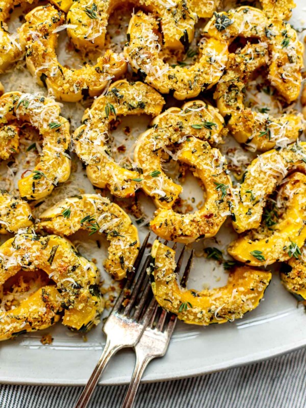 Parmesan Crusted Roasted Delicata Squash arranged on a creamy white serving platter with a dark brown rim. Two forks rest on one side of the platter and the platter sits atop a gray and white striped linen napkin.