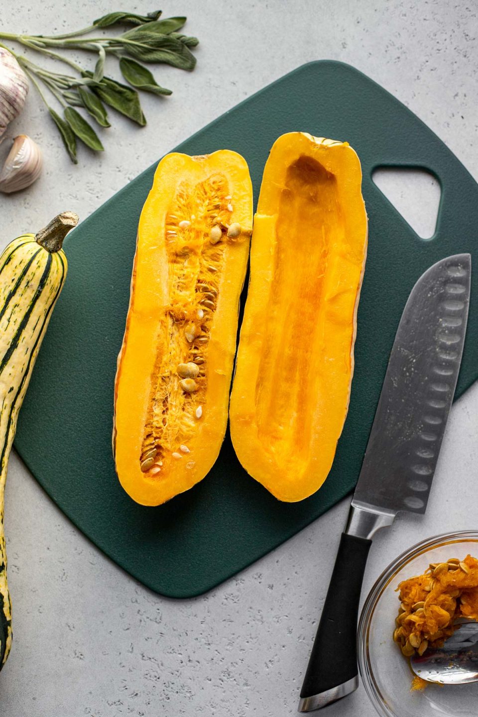 Two cut halves of a delicata squash lay skin side down on a dark green plastic cutting board. One of the halves has had the seeds scooped out, while the seeds remain intact in the other. A chef's knife rests on the cutting board. A small clear glass bowl filled with delicata seeds and a spoon, another whole delicata squash, some loose fresh herbs, and garlic surround the cutting board.