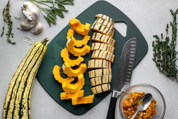 Cut slices of delicata squash arranged on a dark green plastic cutting board. A chef's knife rests on the cutting board. A small clear glass bowl filled with delicata seeds and a spoon, another whole delicata squash, some loose fresh herbs, and garlic surround the cutting board.