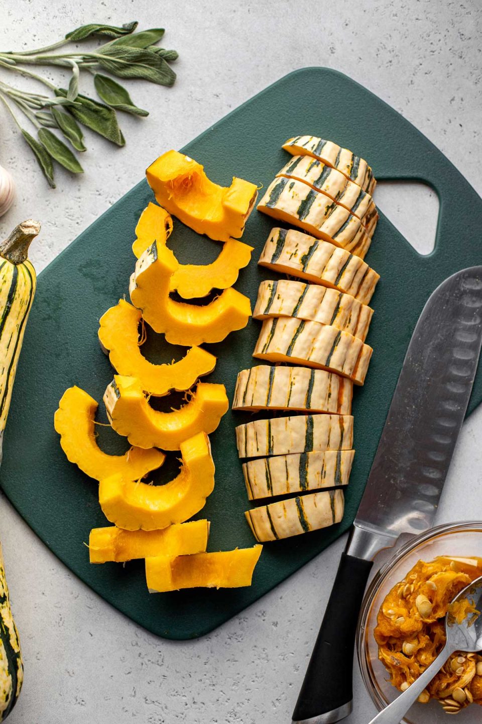 Cut slices of delicata squash arranged on a dark green plastic cutting board. A chef's knife rests on the cutting board. A small clear glass bowl filled with delicata seeds and a spoon, another whole delicata squash, and some loose fresh herbs surround the cutting board.