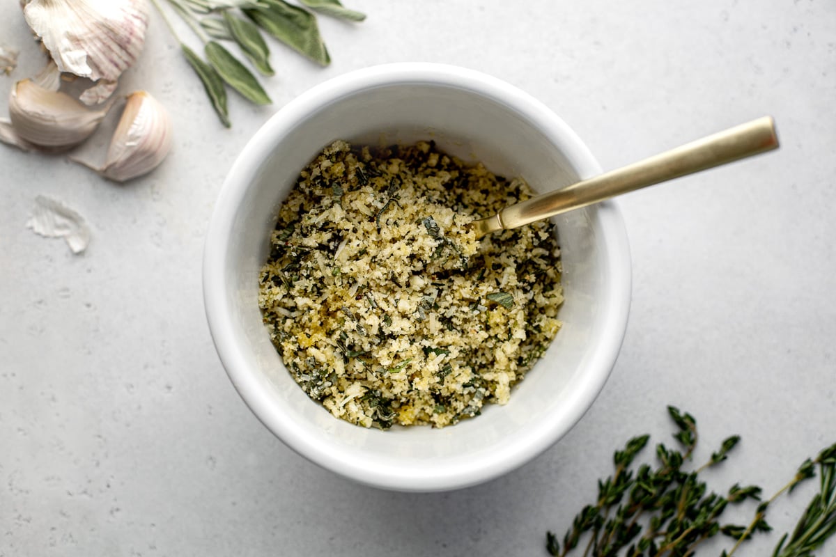 A small white bowl filled with a herbed panko breadcrumb mixture sits atop a creamy white textured surface. A gold spoon rests inside of the bowl and loose sprigs of fresh herbs and whole garlic cloves surround the bowl.