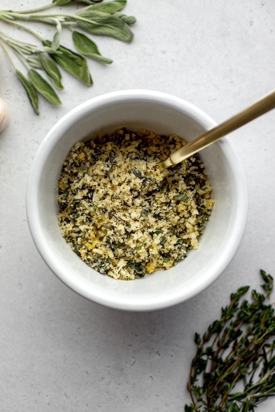 A small white bowl filled with a herbed panko breadcrumb mixture sits atop a creamy white textured surface. A gold spoon rests inside of the bowl and loose sprigs of fresh herbs surround the bowl.