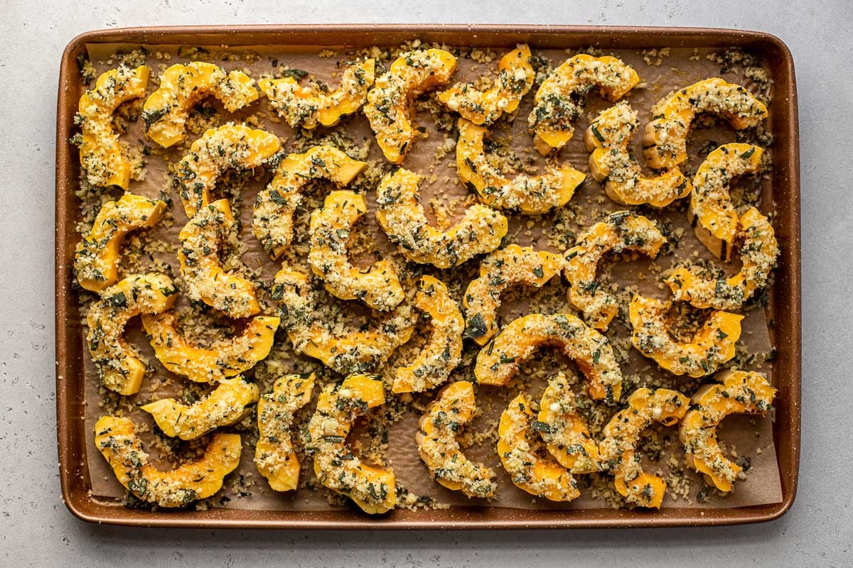 Slices of Delicata Squash seasoned with parmesan cheese, panko breadcrumbs, and fresh herbs are arranged on a baking sheet lined with brown parchment paper for roasting. The baking sheet ​sits atop a creamy white textured surface