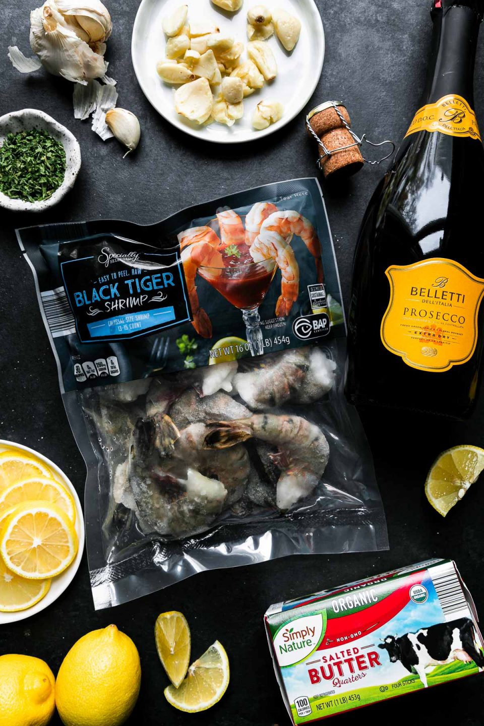 Prosecco Butter Poached Shrimp ingredients arranged on a black textured surface: Specially Selected Black Tiger Shrimp, Belletti Prosecco, Simply Nature Organic Salted Butter, lemon, garlic, Stonemill Parsley Flakes.