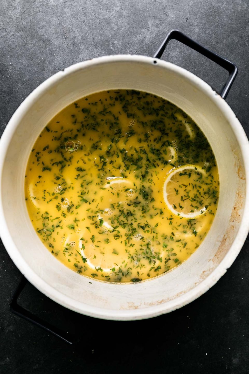 How to Make Prosecco Butter Poached Shrimp, Step 3: Preparing the Prosecco Butter Poaching Liquid. Reduced Prosecco combined with butter, lemon juice, sliced lemon, crushed garlic cloves and dried parsley fills a medium saucepan. The saucepan sits atop a black textured surface.