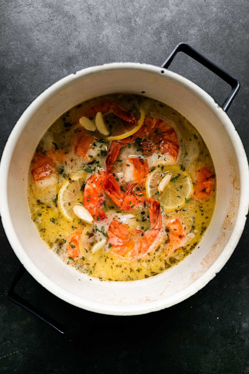 How to Make Prosecco Butter Poached Shrimp, Step 4: Poach the shrimp. Gently poached shrimp rest inside of a saucepan filled with a lemon garlic Prosecco butter poaching liquid. The saucepan sits atop a black textured surface and a gray and white striped napkin rests alongside the saucepan.
