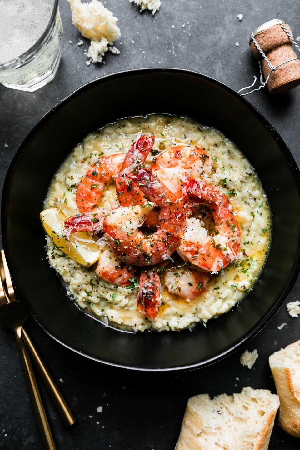 An overhead shot of Prosecco Butter Poached Shrimp served atop a bed of risotto in a black bowl rests atop a black textured surface. A few spoonfuls of poaching liquid have been poured over the shrimp & the dish has been garnished with freshly grated parmesan cheese and a lemon wedge tucked alongside the shrimp. Gold silverware, a few pieces of loose crusty bread for dipping, bits of breadcrumbs, a glass filled with Prosecco, and a Prosecco bottle cork surround the bowl of shrimp.