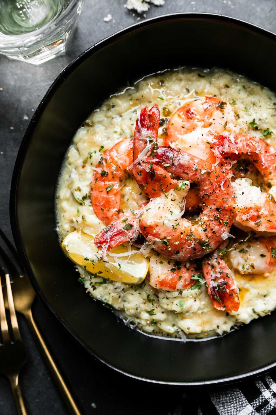 An overhead shot of Prosecco Butter Poached Shrimp served atop a bed of risotto in a black bowl rests atop a black textured surface. A few spoonfuls of poaching liquid have been poured over the shrimp & the dish has been garnished with freshly grated parmesan cheese and a lemon wedge tucked alongside the shrimp. Gold silverware, a glass filled with Prosecco, and a black and white plaid linen napkin surround the bowl of shrimp.