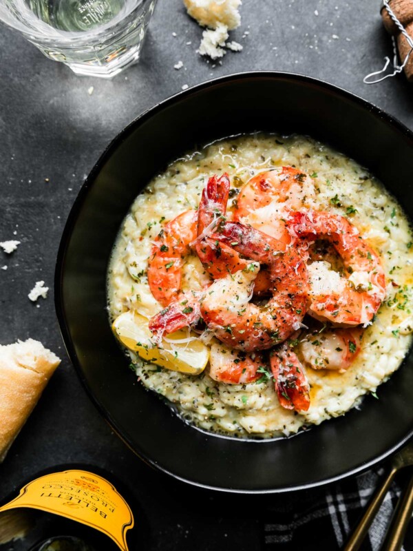 An overhead shot of Prosecco Butter Poached Shrimp served atop a bed of risotto in a black bowl rests atop a black textured surface. A few spoonfuls of poaching liquid have been poured over the shrimp & the dish has been garnished with freshly grated parmesan cheese and a lemon wedge tucked alongside the shrimp. Gold silverware, a bottle of Prosecco, a glass filled with Prosecco, pieces of crusty bread and crumbs, a Prosecco bottle cork, and a black and white plaid linen napkin surround the bowl of shrimp.