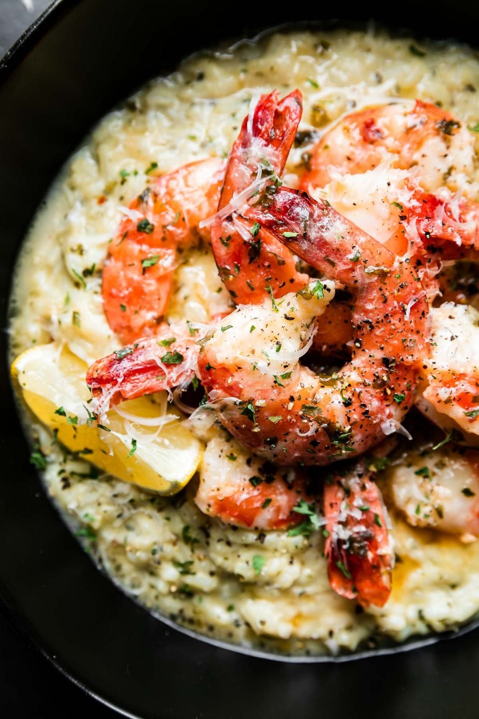 A close up overhead shot of Prosecco Butter Poached Shrimp served atop a bed of risotto. A few spoonfuls of poaching liquid have been poured over the shrimp & the dish has been garnished with freshly grated parmesan cheese and a lemon wedge tucked alongside the shrimp.
