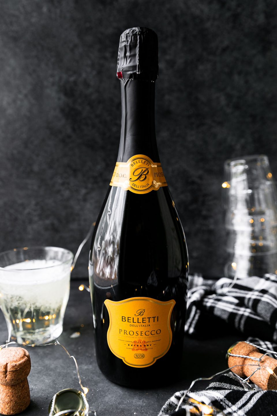An unopened bottle of Belletti Prosecco from ALDI sits atop a black textured surface in front of a black textured background. A few opened corks, a string of twinkling lights, a clear glass filled with Prosecco, a black and white plaid linen napkin, and a stack of upside-down glasses surround the bottle.
