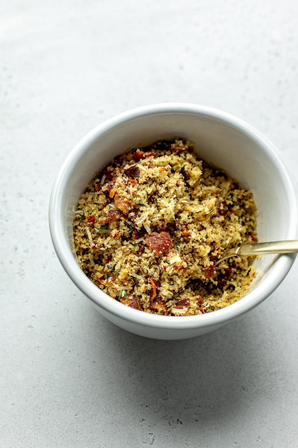 A side angle shot of a small white ceramic bowl filled with bacon breadcrumb topping and a gold spoon rests atop a creamy white textured surface.