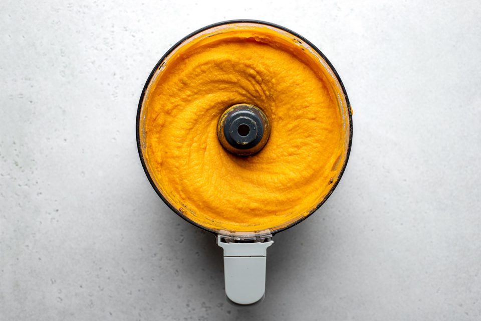 An overhead shot of a food processor bowl filled with pureed sweet potatoes. The food processor bowl sits atop a creamy white textured surface.