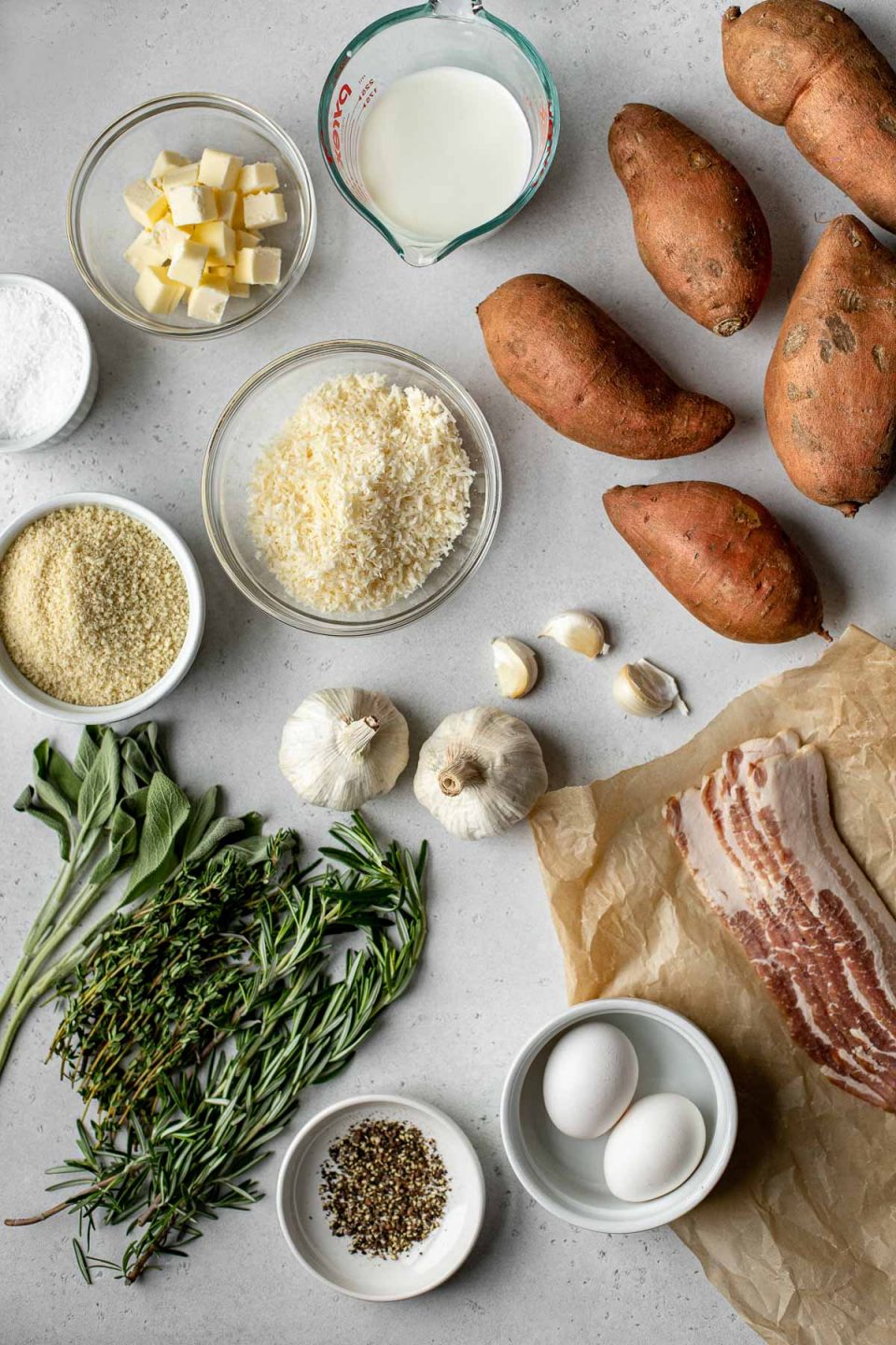 Savory Sweet Potato Casserole ingredients arranged on a creamy white textured surface: sweet potatoes, garlic, unsalted butter, whole milk, grated parmesan, eggs, kosher salt, ground black pepper, bacon, fresh herbs - rosemary, sage, thyme, panko breadcrumbs.