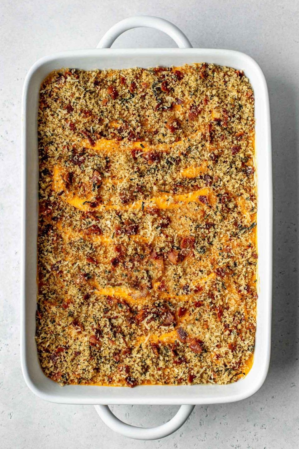 An overhead shot of a baking dish filled with assembled and baked Savory Sweet Potato Casserole topped with bacon breadcrumb topping. The baking dish sits atop a creamy white textured surface.