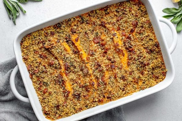 An overhead shot of a baking dish filled with assembled and baked Savory Sweet Potato Casserole topped with bacon breadcrumb topping. The baking dish sits atop a creamy white textured surface. A gray and white striped linen napkin is tucked underneath the baking dish and a few loose fresh herb and a parmesan rind surround the baking dish.