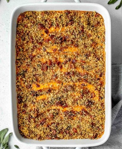 An overhead shot of a baking dish filled with assembled and baked Savory Sweet Potato Casserole topped with bacon breadcrumb topping. The baking dish sits atop a creamy white textured surface. A gray and white striped linen napkin is tucked underneath the baking dish. A few loose fresh herbs and a parmesan rind surround the baking dish.