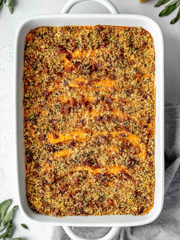 An overhead shot of a baking dish filled with assembled and baked Savory Sweet Potato Casserole topped with bacon breadcrumb topping. The baking dish sits atop a creamy white textured surface. A gray and white striped linen napkin is tucked underneath the baking dish. A few loose fresh herbs and a parmesan rind surround the baking dish.