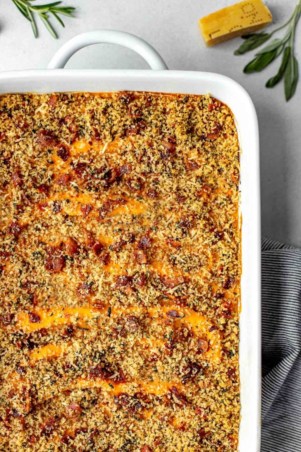 An overhead shot of a baking dish filled with assembled and baked Savory Sweet Potato Casserole topped with bacon breadcrumb topping. The baking dish sits atop a creamy white textured surface. A gray and white striped linen napkin is tucked underneath the baking dish. A few loose fresh herbs and a parmesan rind surround the baking dish.