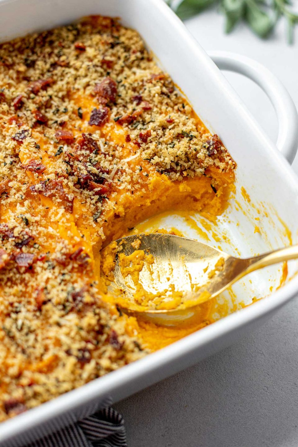 A side angle shot of a white baking dish filled with assembled and baked Savory Sweet Potato Casserole topped with bacon breadcrumb topping. A spoon rests inside of the baking dish with a portion of the casserole scooped out. The baking dish sits atop a creamy white textured surface. A gray and white striped linen napkin is tucked underneath the baking dish. A few loose fresh herbs surround the baking dish in the background.