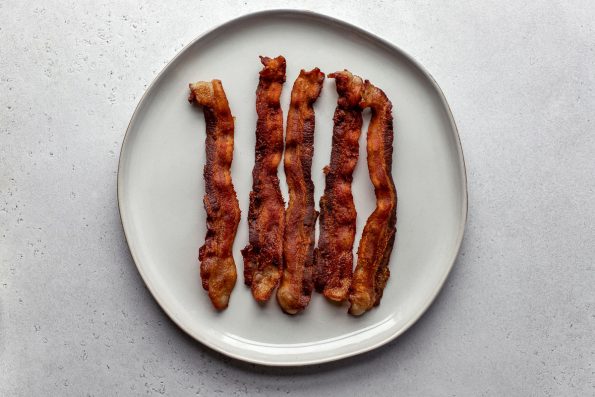 Five slices of crispy cooked bacon rest atop a white ceramic plate that sits atop a creamy white textured surface.