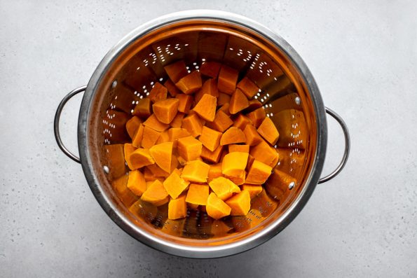 Boiled sweet potatoes strained in a stainless steel colander. The colander rests atop a creamy white textured surface.