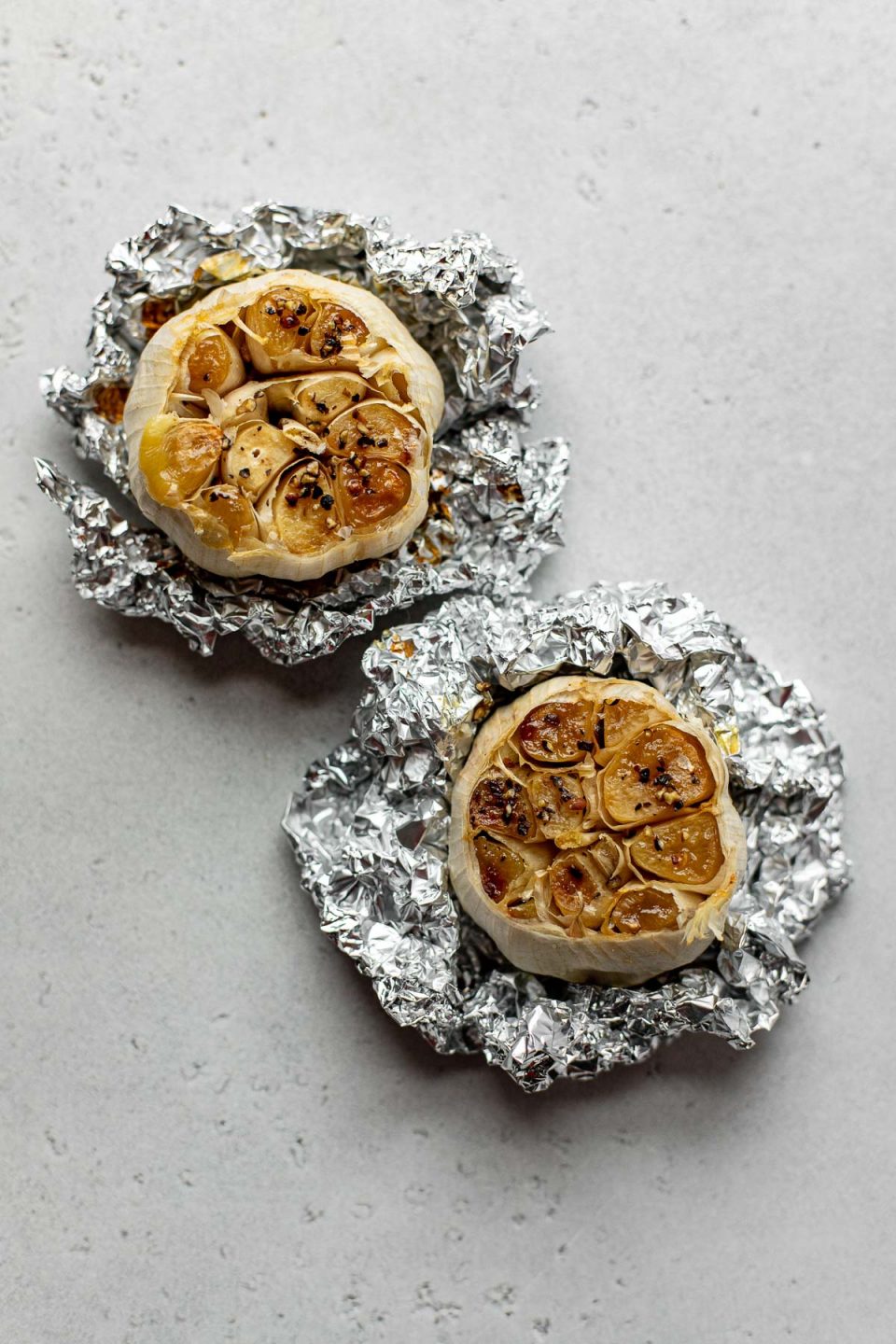 Two heads of roasted garlic with their tops cut off are seasoned with olive oil, kosher salt and pepper rest atop sheets of crumpled aluminum foil. Each bundle of roasted garlic sits atop a creamy white textured surface.