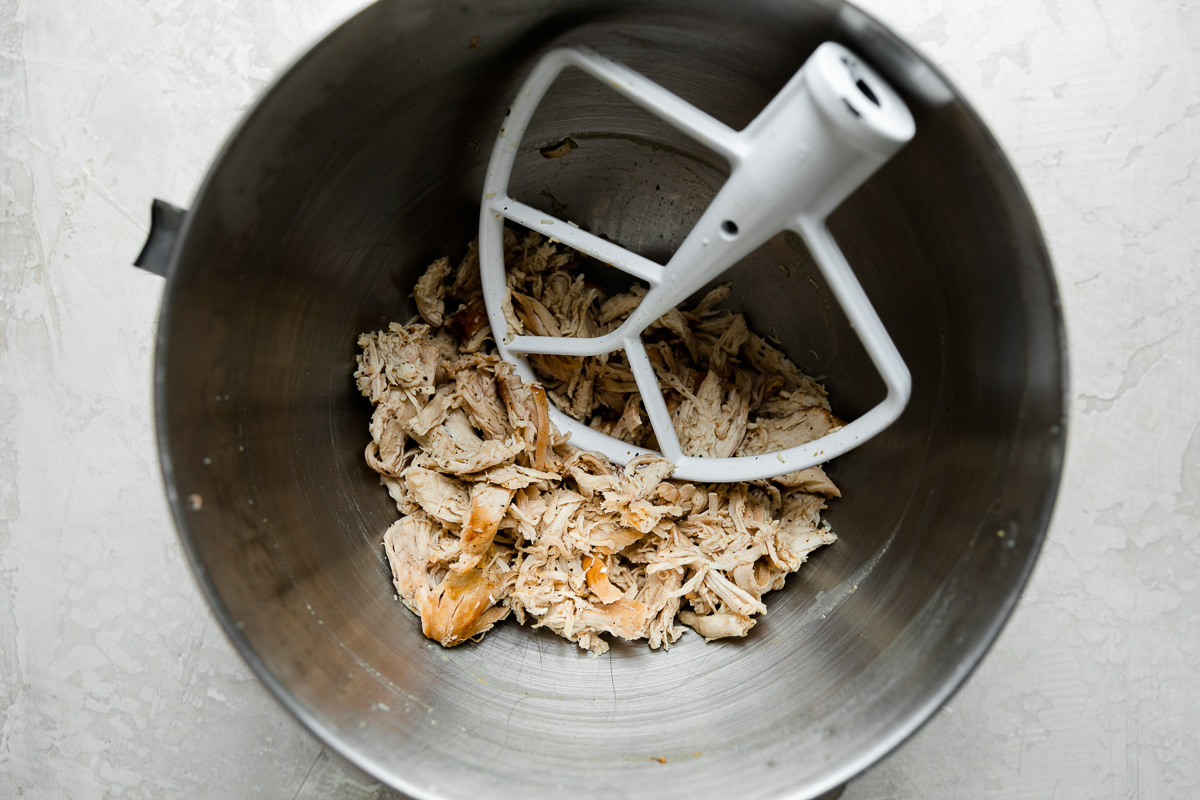 Three browned chicken breasts, shredded in a stand mixer bowl. The paddle attachment for the stand mixer rests inside of the bowl and the bowl sits atop a creamy white textured surface.