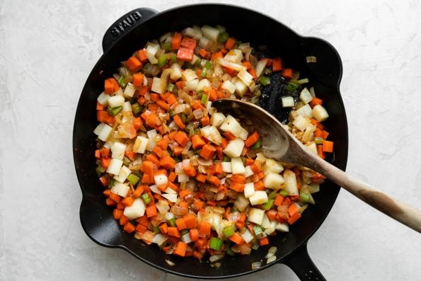 A combination of diced carrots, celery, onion, and aromatics brown in a black Staub cast iron skillet. The skillet sits atop a creamy white textured surface and a wooden spoon rests inside the skillet.