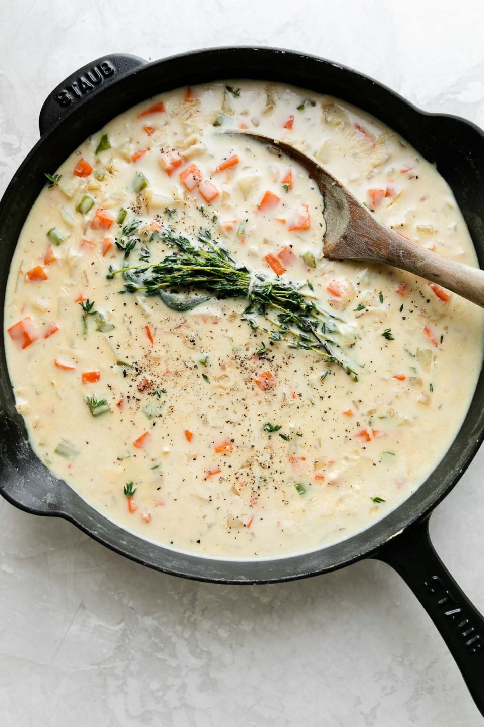 A black Staub cast iron skillet is filled with sauce to make Skillet Chicken Pot Pie. Fresh rosemary, thyme, and sage tied together with kitchen twine has been added to the sauce and it has also been seasoned with kosher salt and ground black pepper. The skillet sits atop a creamy white textured surface and a wooden spoon rests inside the skillet.