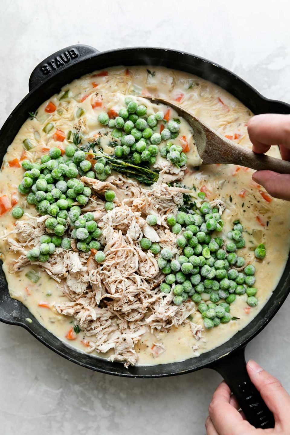 A black Staub cast iron skillet is filled with sauce to make Skillet Chicken Pot Pie. Frozen peas and shredded chicken has been added to the sauce to assemble the pot pie filling. The skillet sits atop a creamy white textured surface. A woman's hands hold the skillet handle while her other hand holds a wooden spoon using it to mix the pot pie filling.