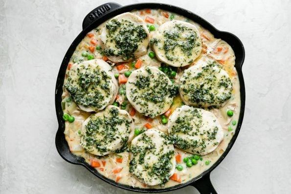 An overhead shot of a black Staub cast iron skillet filled Skillet Chicken Pot Pie filling topped with uncooked extra flaky biscuits arranged over top. Each biscuits has been brushed with a herb butter and the skillet sits atop a creamy white textured surface.