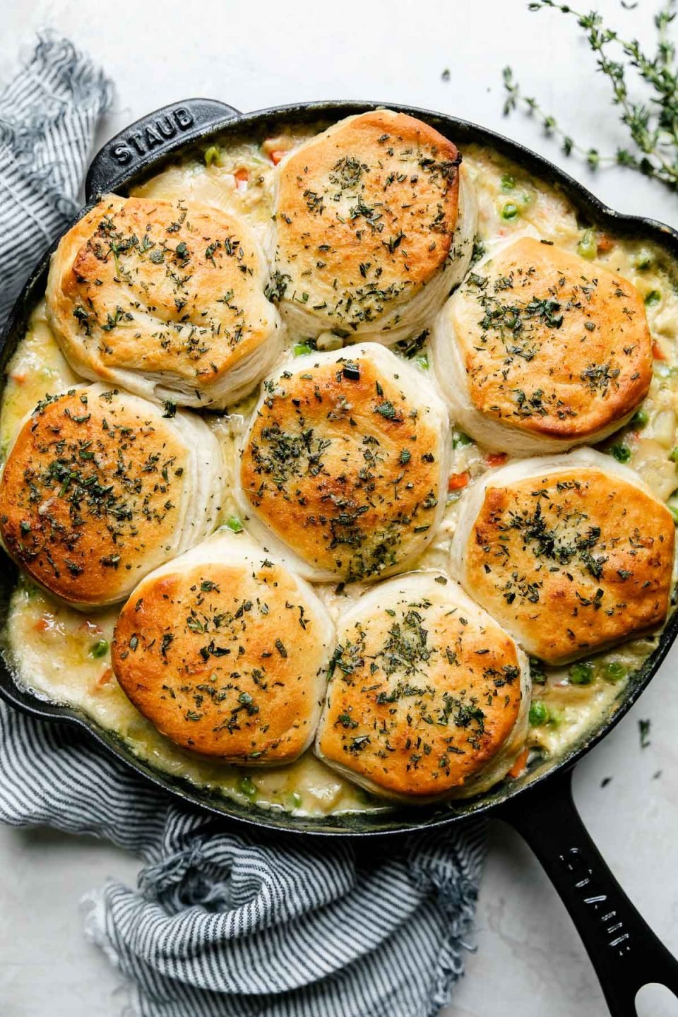 An overhead shot of Skillet Chicken Pot Pie with Biscuits in a black Staub cast iron skillet that sits atop a creamy white textured surface. A blue and white striped linen napkin rests alongside the skillet and sprigs of fresh thyme also surround the skillet.