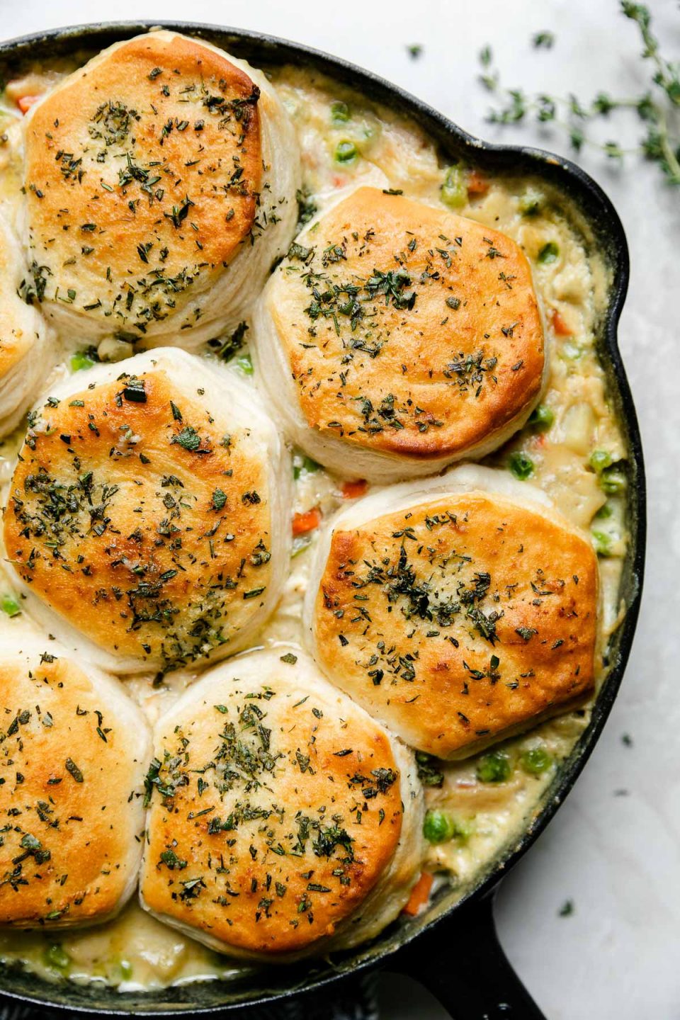 An overhead shot of Skillet Chicken Pot Pie with Biscuits in a black Staub cast iron skillet that sits atop a creamy white textured surface. Sprigs of fresh thyme surround the skillet.