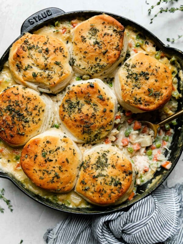 An overhead shot of Skillet Chicken Pot Pie with Biscuits in a black Staub cast iron skillet that sits atop a creamy white textured surface. One serving of the pot pie & one of the biscuits has been removed from the skillet. A gold spoon rests inside of the skillet. A blue and white striped linen napkin is tied around the skillet handle while sprigs of fresh thyme surround the skillet.