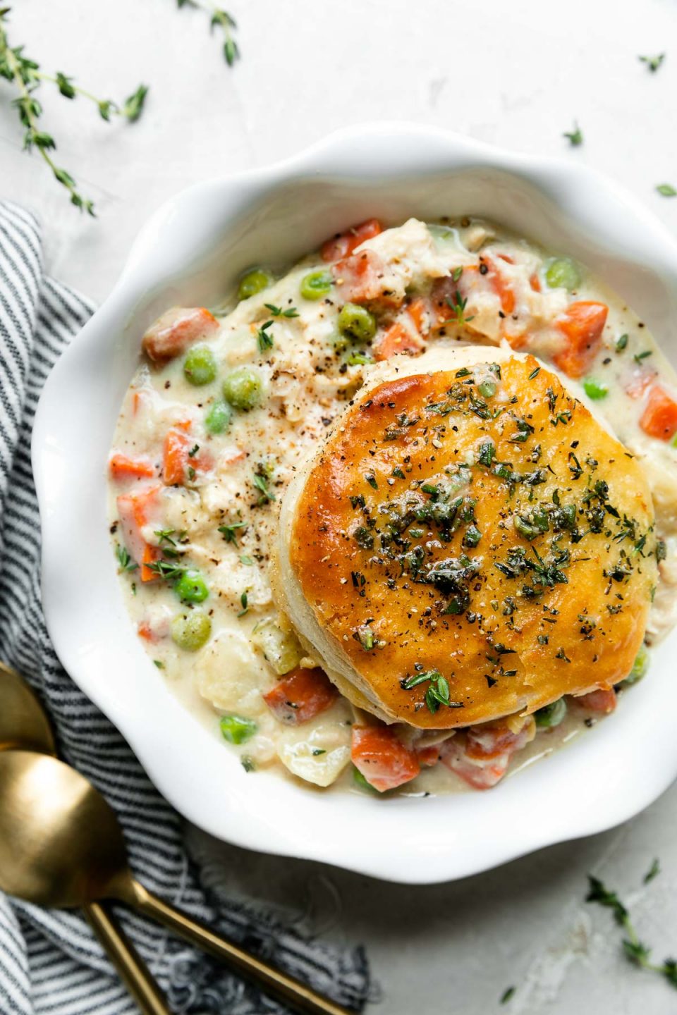 An overhead shot of Skillet Chicken Pot Pie with Biscuits served on a white plate with scalloped edges that rest atop a creamy white textured surface. Two gold spoons, a blue and white striped linen napkin, and a few sprigs of fresh thyme surround the plate.
