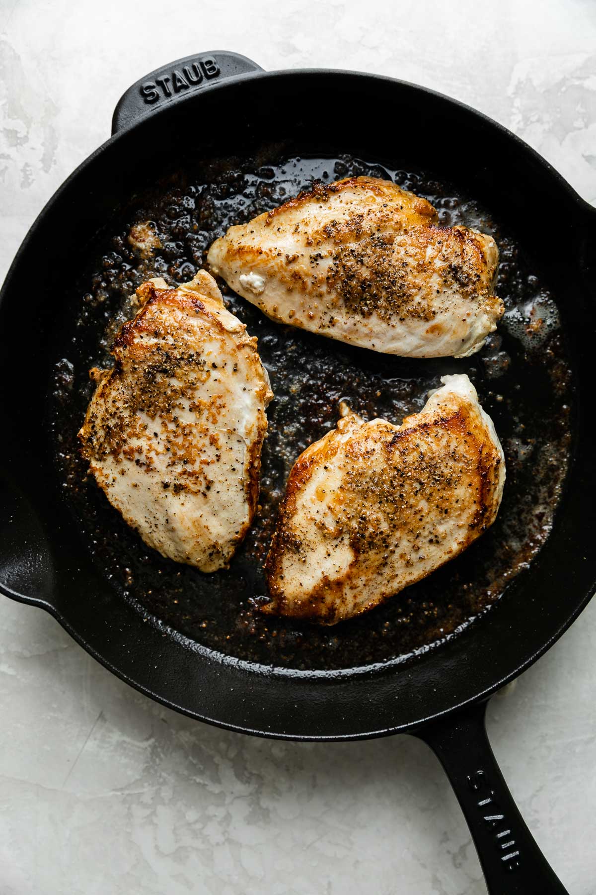 Three browned chicken breasts, seasoned with salt and pepper, in a Staub black cast iron skillet resting atop a creamy white textured surface.