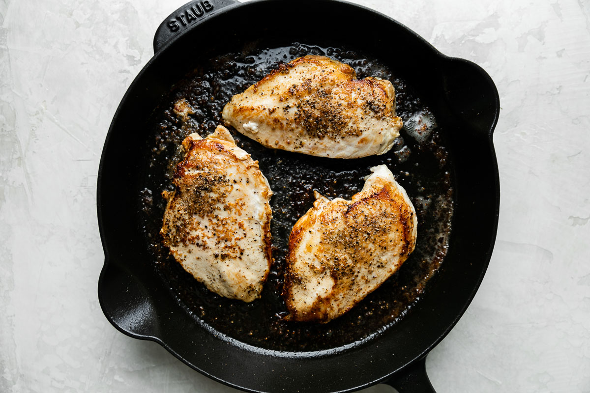 Three browned chicken breasts, seasoned with salt & pepper, in a Staub black cast iron skillet rest atop a creamy white textured surface.