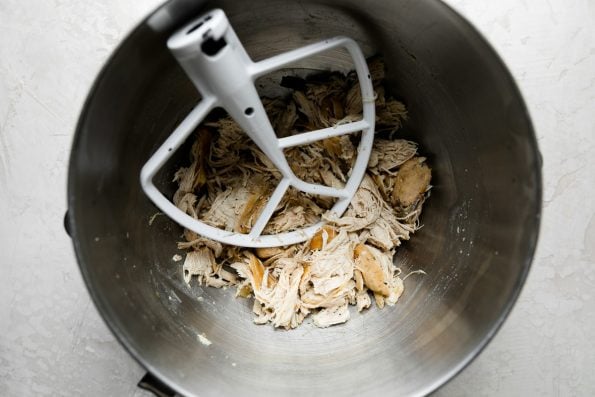 Three browned chicken breasts, shredded in a stand mixer bowl. The paddle attachment for the stand mixer rests inside of the bowl and the bowl sits atop a creamy white textured surface.