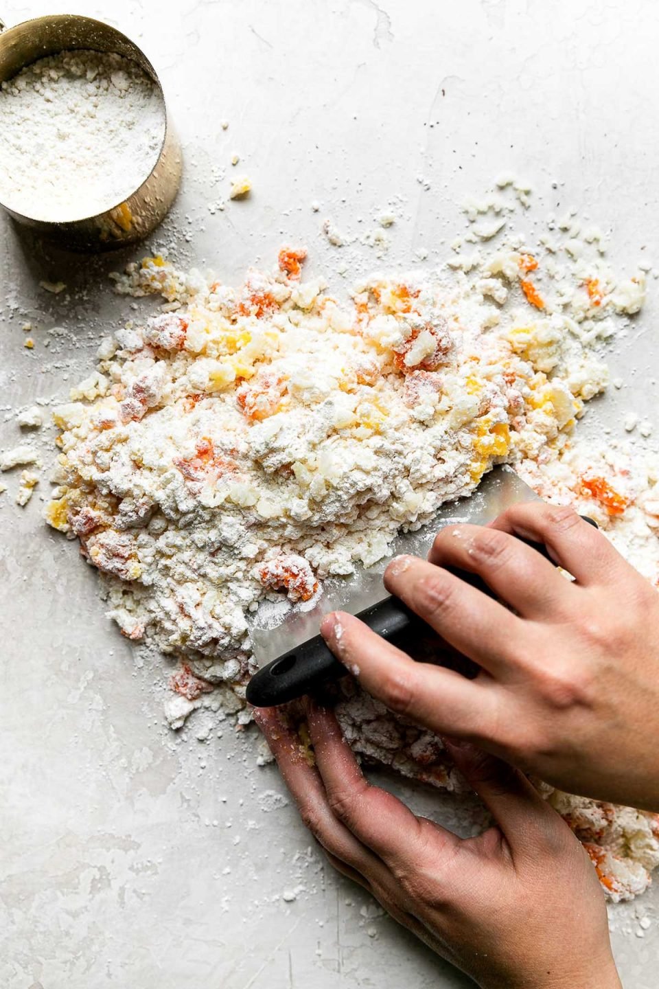 A woman's hands hold a bench scraper and use it to combine the components of the homemade sweet potato gnocchi dough. The bench scraper helps to cut the flour into the potato mixture. The dough forms on a creamy white plaster textured surface and a gold measuring cup filled with extra pastry flour rests alongside the mixture.