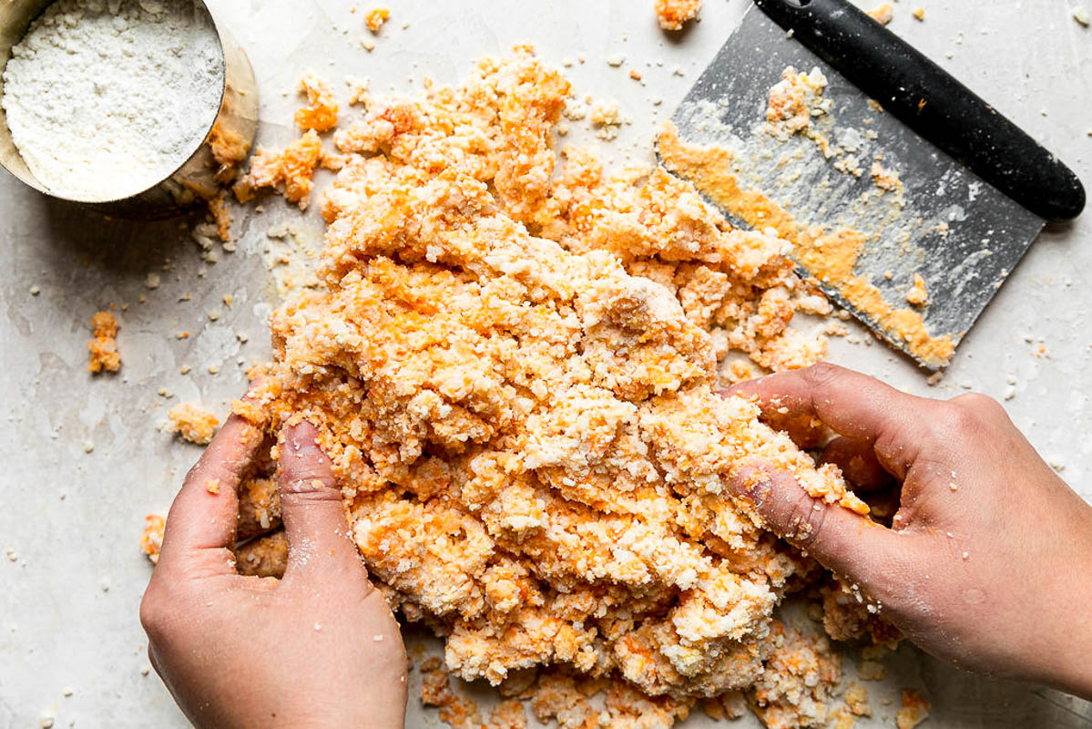 A woman's hands mix together homemade sweet potato gnocchi dough. The dough has begun to form on a creamy white plaster textured surface and a gold measuring cup filled with extra pastry flour rests alongside the mixture. A bench scraper used to help cut in the flour also rests alongside the dough mixture.