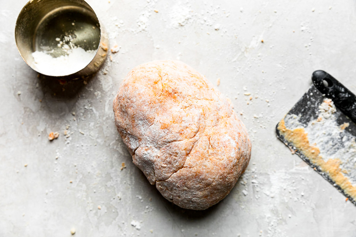 A dough ball of homemade sweet potato gnocchi sits atop a white plaster textured surface with a gold measuring cup filled with extra pastry flour and a bench scraper used to help cut flour into the dough rests alongside the formed dough ball.