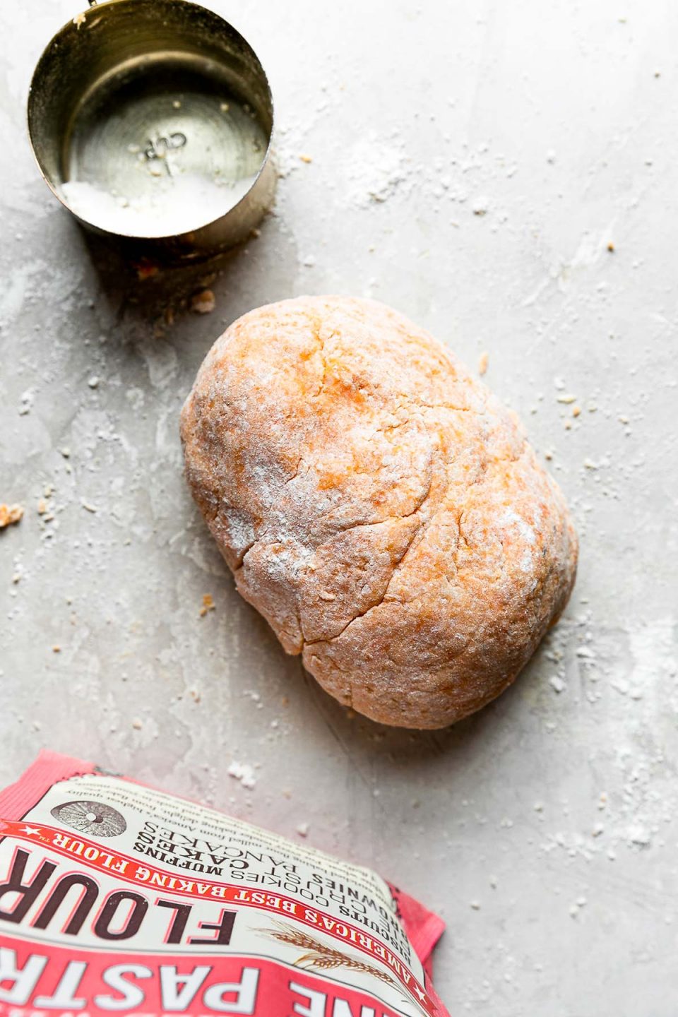 A dough ball of homemade sweet potato gnocchi sits atop a white plaster textured surface dusted with white pastry flour. A gold measuring cup filled with extra pastry flour and a bag of Bob's Red Mill Unbleached White Fine Pastry Flour sits upright alongside the formed dough ball.