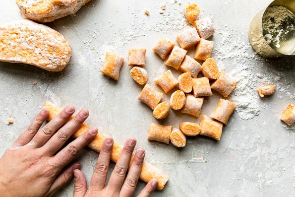 A woman's hands work to roll out cut sections of fresh homemade sweet potato gnocchi dough into long uniform pieces of dough. Approximately 1-inch long pieces of gnocchi dough about 1/2-inch thick have been cut and are arranged in a pile while other sections of gnocchi dough wait to be rolled out. The process takes place on a creamy white textured surface. A gold measuring cup filled with extra pastry flour also rests on the surface.