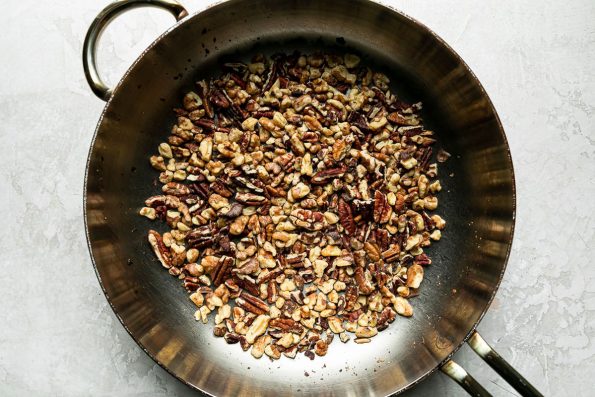 A stainless steel frying pan is filled with chopped walnuts for toasting. The frying pan sits atop a creamy white textured surface.