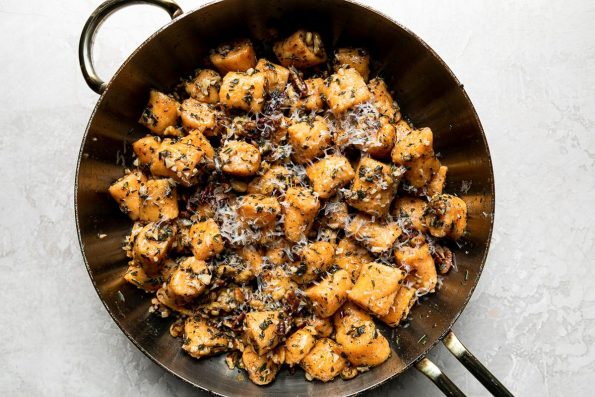 An overhead shot of Sweet Potato Gnocchi with Garlic Herb Brown Butter sauce in a metal sauce pan. The gnocchi is garnished with freshly grated parmesan cheese and the saucepan sits atop a creamy white textured surface.