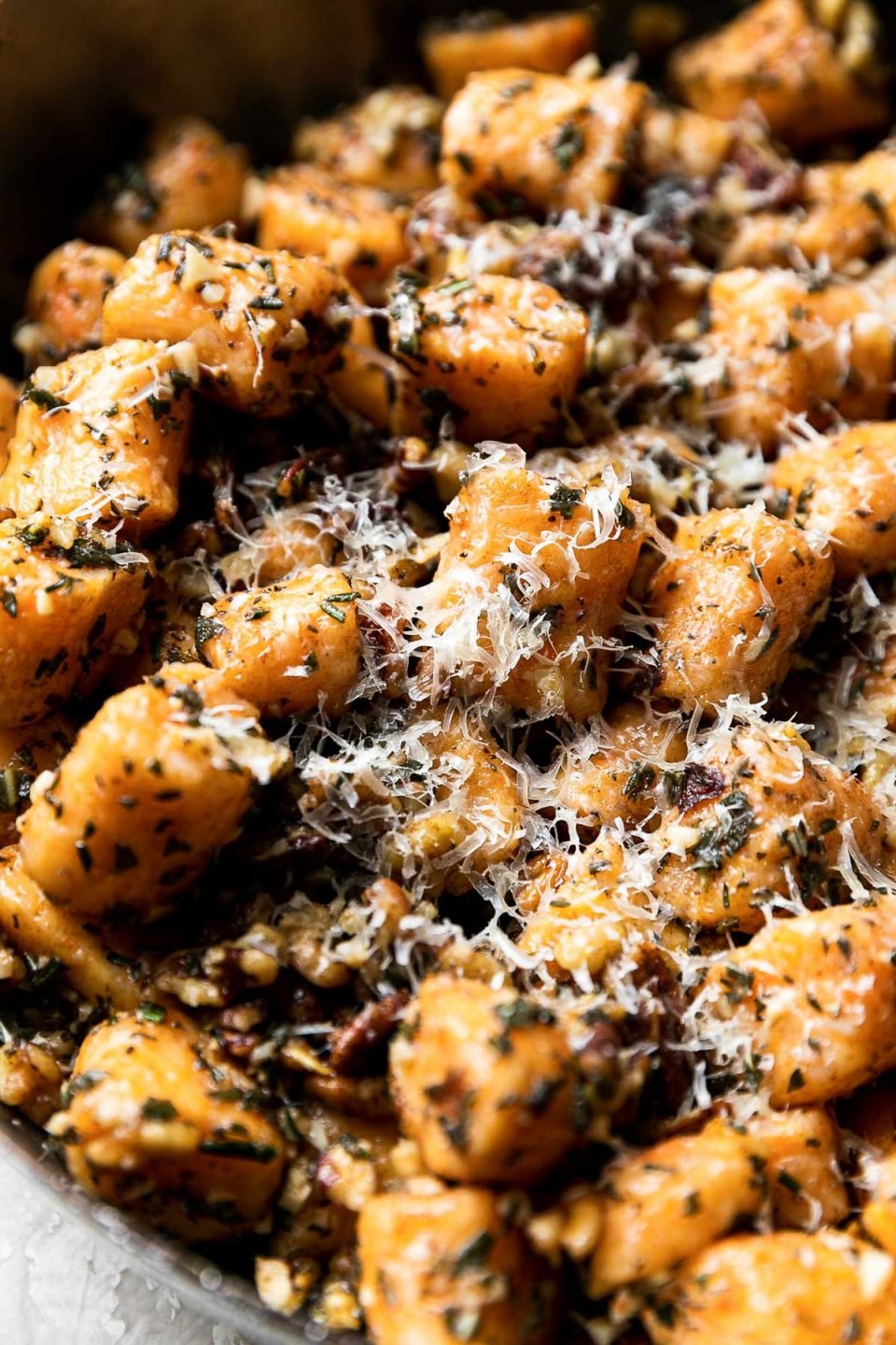 A side angle shot of Sweet Potato Gnocchi with Garlic Herb Brown Butter sauce in a metal sauce pan. The gnocchi is garnished with freshly grated parmesan cheese and the saucepan sits atop a creamy white textured surface.