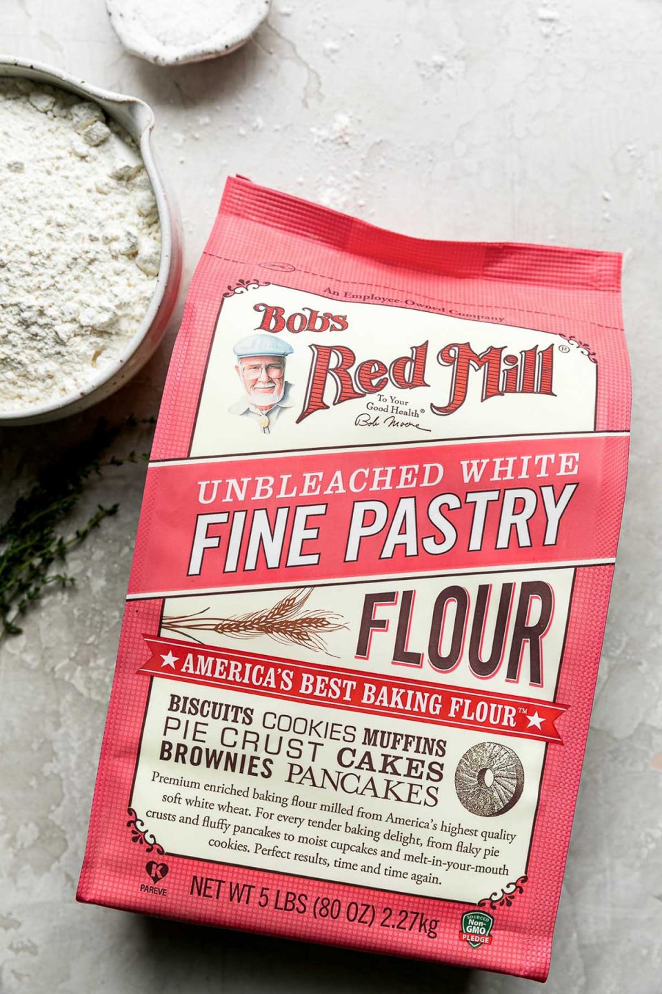 An unopened package of Bob's Red Mill Unbleached White Fine Pastry Flour rests facing upwards on a creamy white plaster textured surface. A small ceramic bowl filled with the pastry flour and a small pinch bowl filled with kosher salt rest alongside of it.